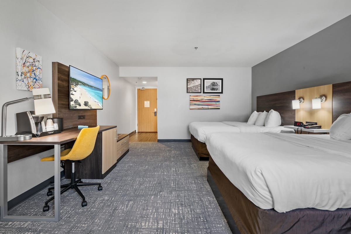 A hotel room with a desk, two beds, a chair, and artwork on the walls.