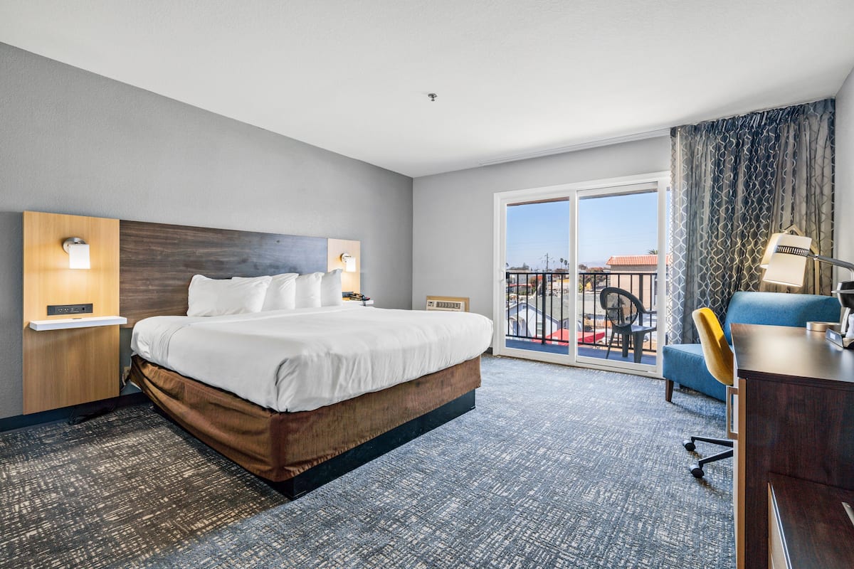 A hotel room with a large bed, a desk, a chair, and a balcony with a view of the city.