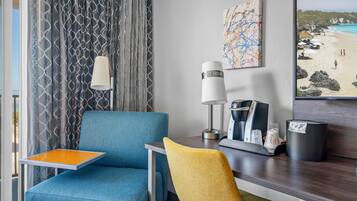 A hotel room with a blue armchair, a yellow chair, a small table with a tray, a lamp, a coffee maker, and a painting of a beach scene.
