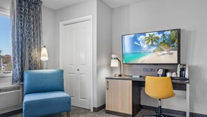 Habitación de hotel con escritorio, un sillón azul, una silla de oficina amarilla, un televisor mostrando una escena de playa y una puerta blanca.