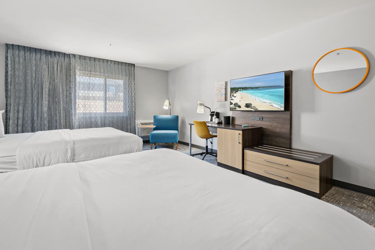A hotel room with two beds, a desk with a chair, a TV, and a large window with curtains.