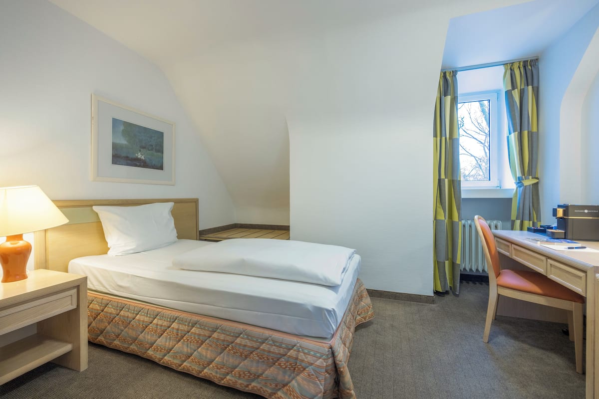 Hotellihuone, jossa on sänky, työpöytä, tuoli, lamppu, seinällä oleva kehystetty kuva ja ikkuna, jossa on verhot.