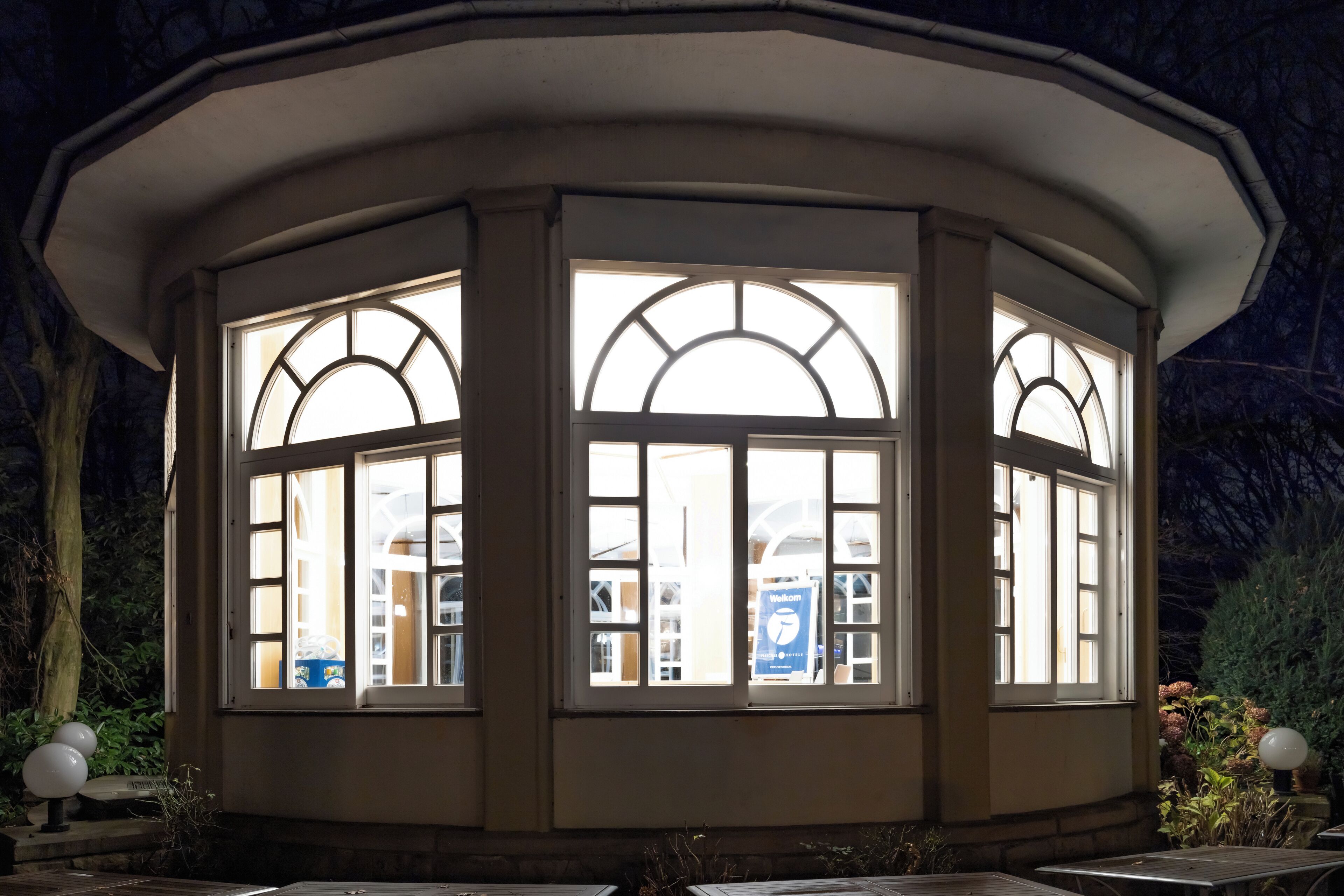 Ein winkelförmiger Pavillon mit großen Fenstern, umgeben von Pflanzen und einer Lampe.