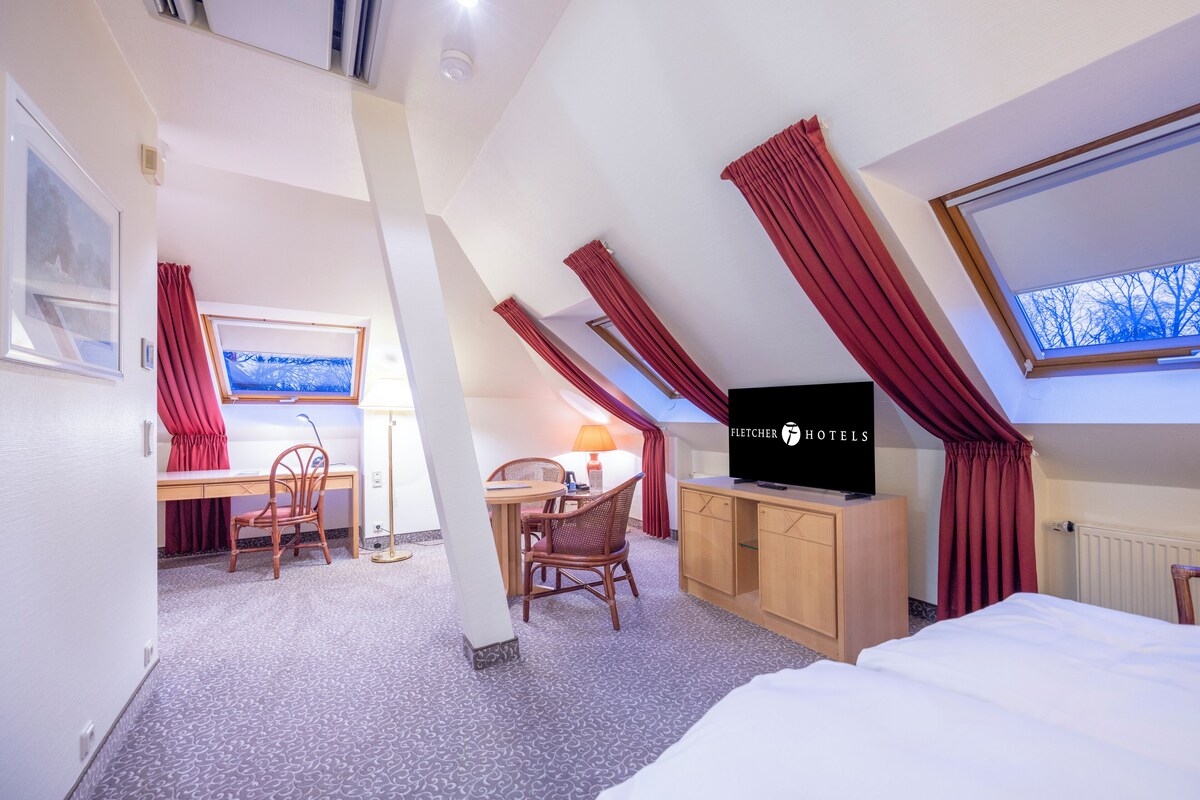 Een hotelkamer met een bed, een bureau met stoel, een televisie en een raam met rode gordijnen.