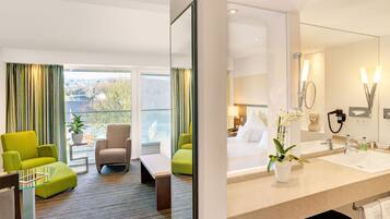 Ein Hotelzimmer mit großem Fenster, einem Bett, einer Sitzecke mit Tisch und Stühlen sowie einem Badezimmer mit Waschbecken und Spiegel.