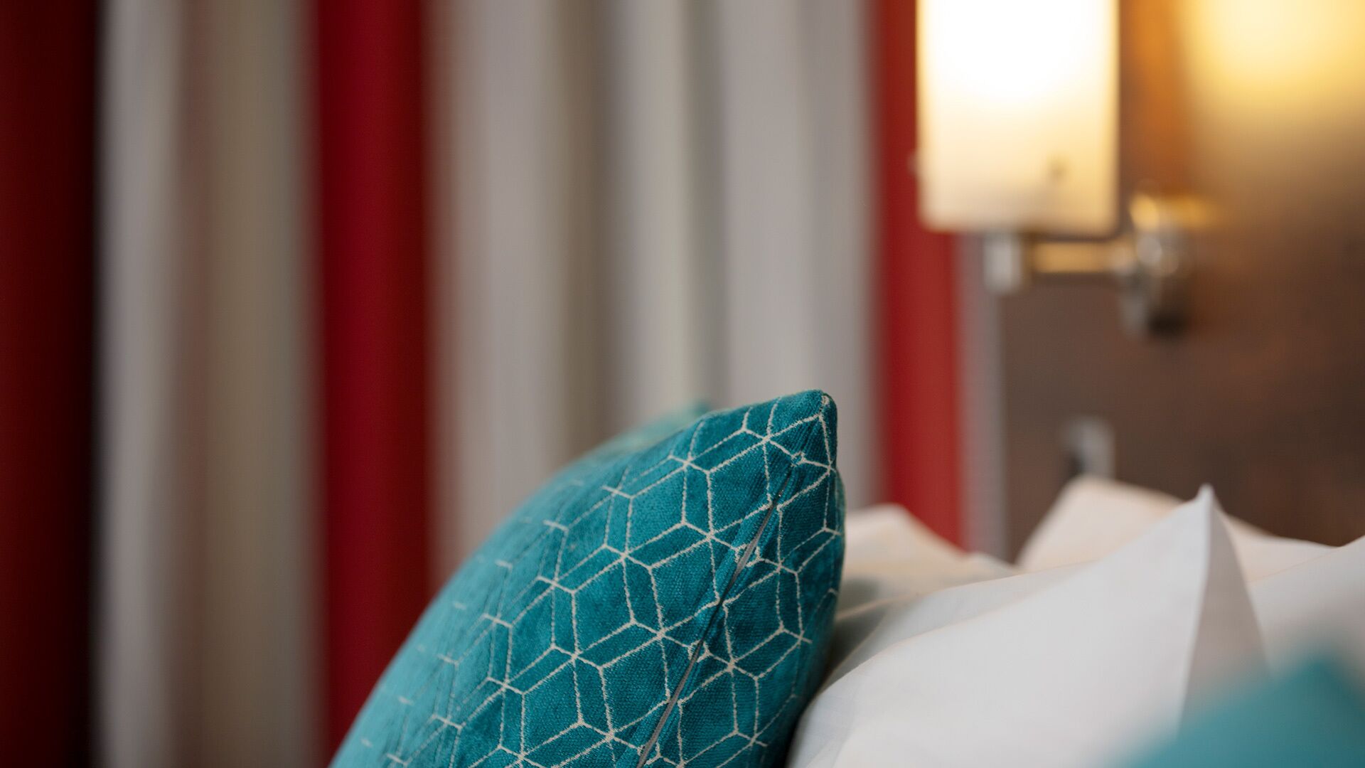 A bed with a teal pillow and white pillow, red curtains, and a wall-mounted light fixture.