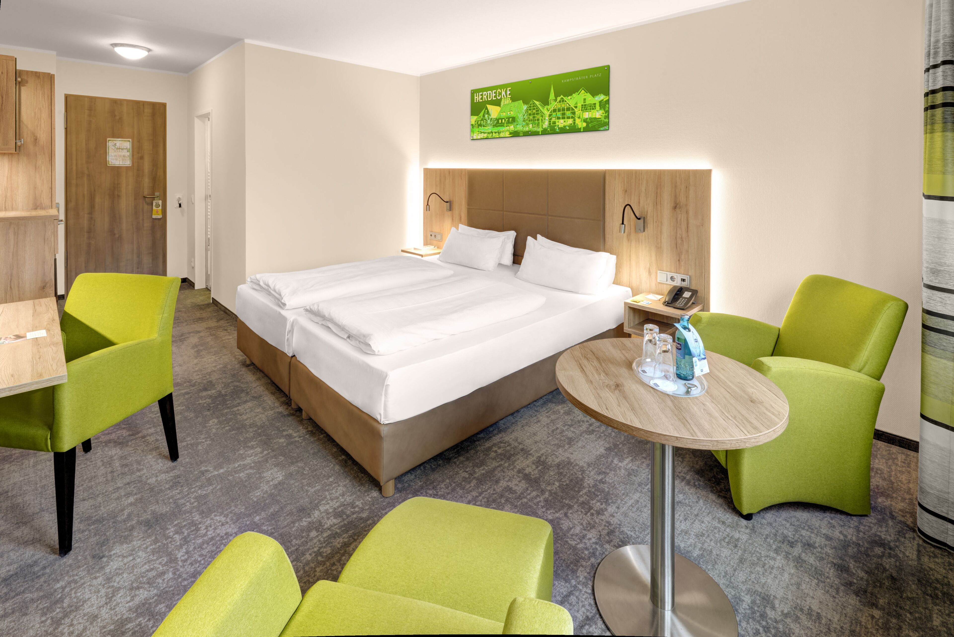 Una habitación de hotel con una cama grande, dos sillones verdes, una mesita redonda y una puerta de madera.