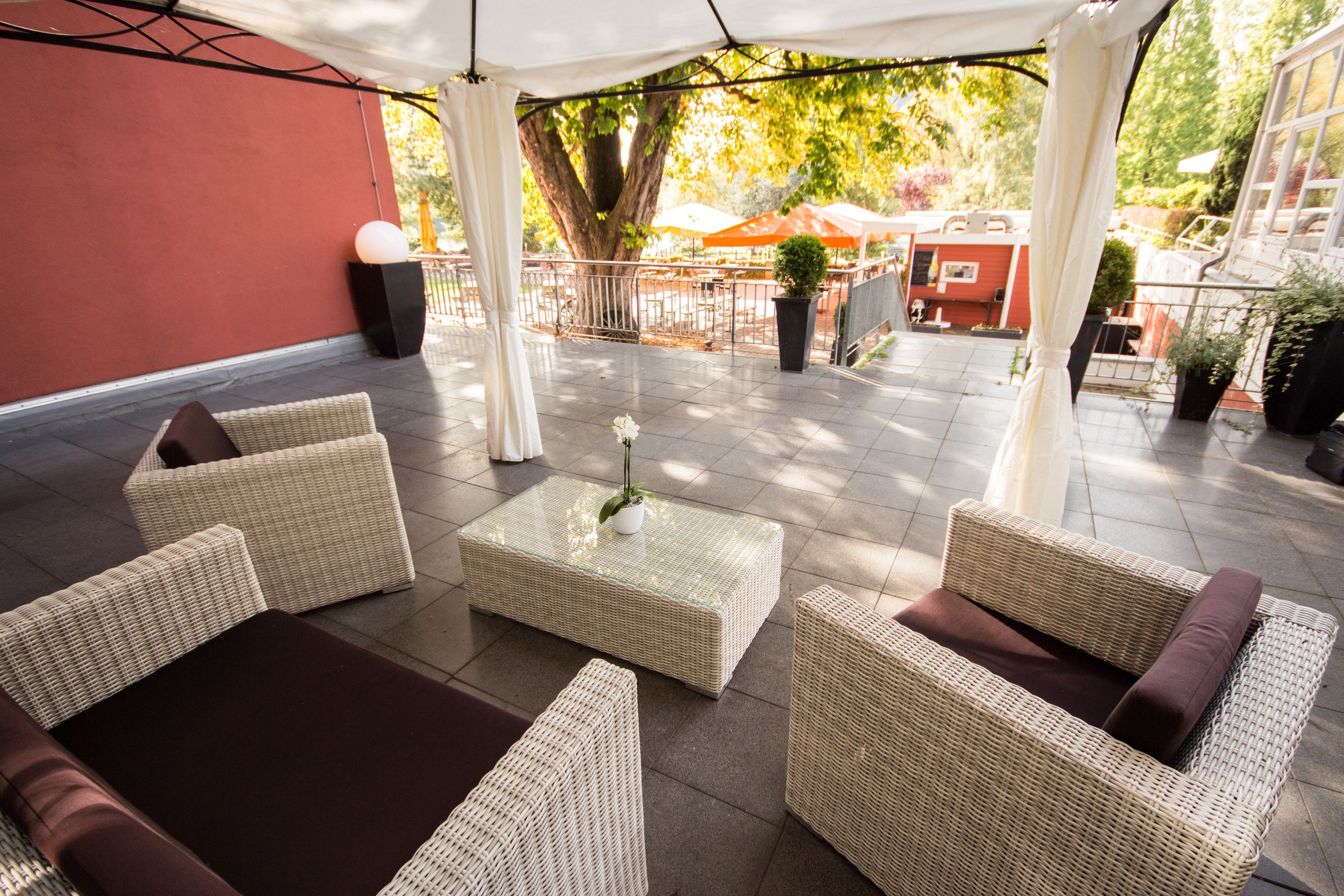 A covered outdoor seating area with wicker furniture and a glass-top table, surrounded by a red wall and potted plants.