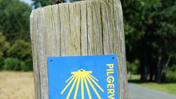 Un poste de madera con un letrero azul que dice "PILGERWEG" y un símbolo de sol amarillo.