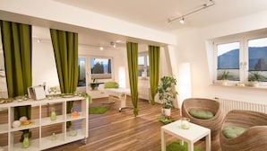 A modern living room with a white sofa, a white coffee table, and a wicker chair. There is a white shelving unit with decorative items and a green curtain.