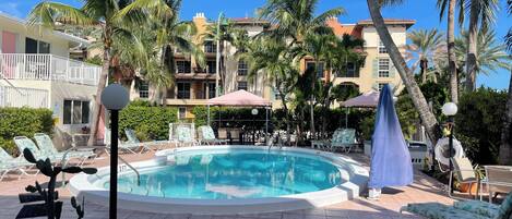 A pool area with lounge chairs, palm trees, and a building with balconies in the background.