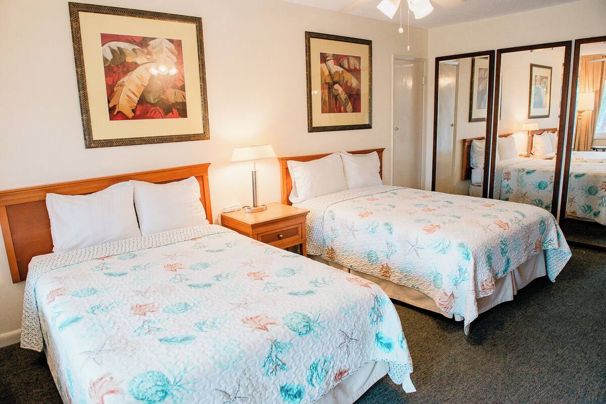 A hotel room with two beds, each with floral bedding, a bedside table with a lamp, and framed artwork on the walls.