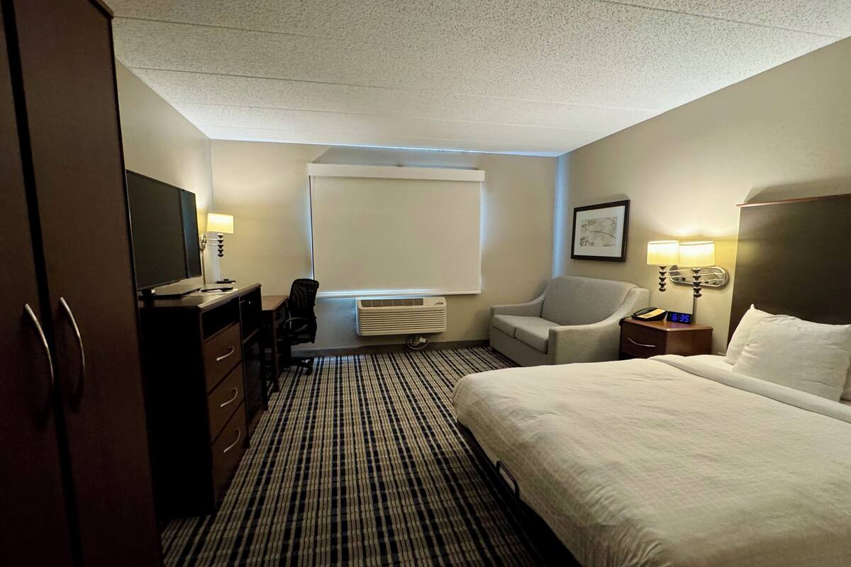 Una habitación de hotel con una cama, un escritorio con silla, televisor, ventana con persianas, aire acondicionado instalado en la pared y una mesita de noche con lámpara.