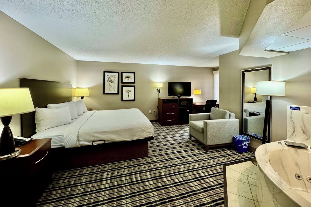 Habitación de hotel con cama, escritorio, silla, televisor, espejo y baño con bañera.