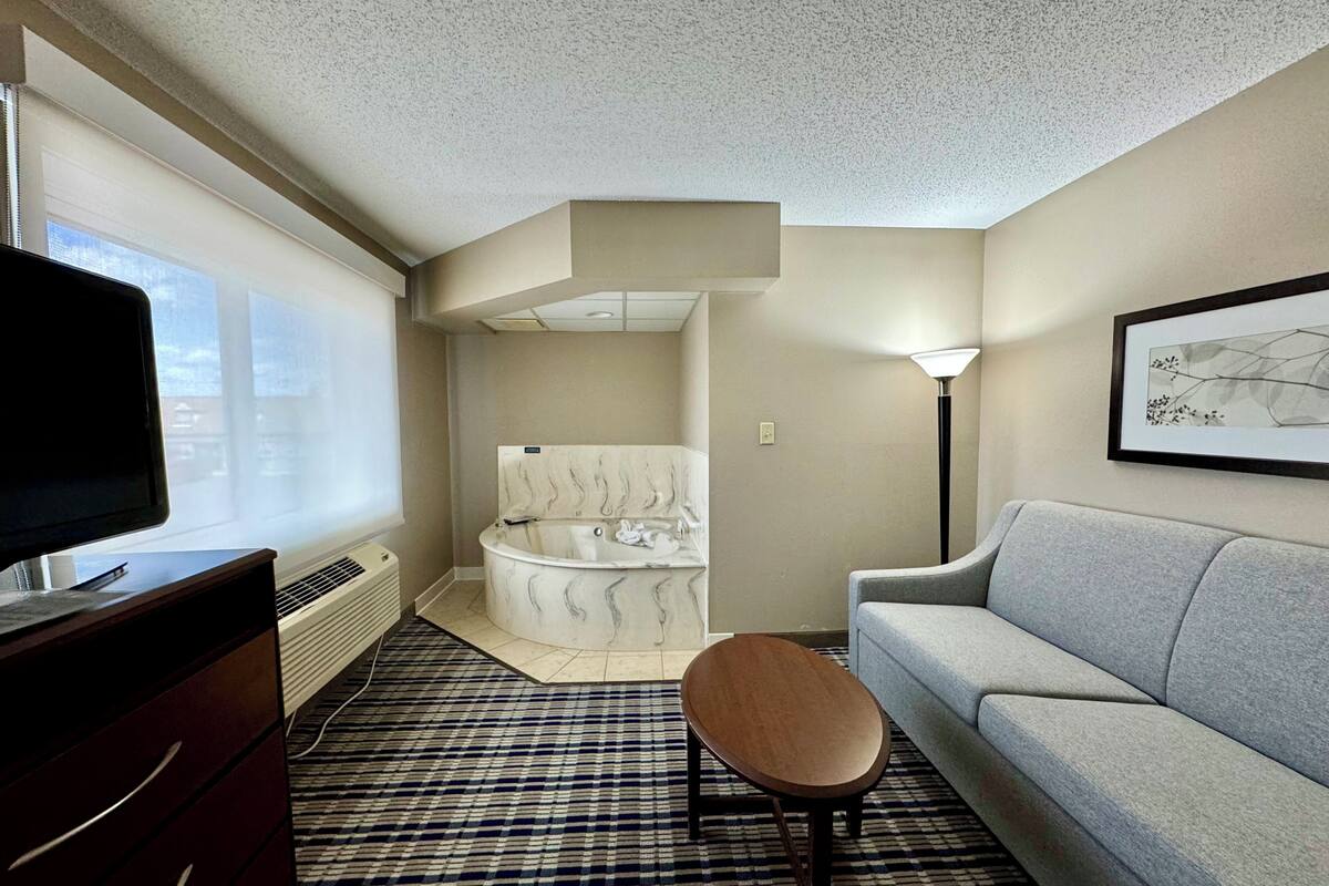 Habitación de hotel con televisor de pantalla plana, sofá, taburete de madera, alfombra estampada, bañera y un cuadro enmarcado en la pared.