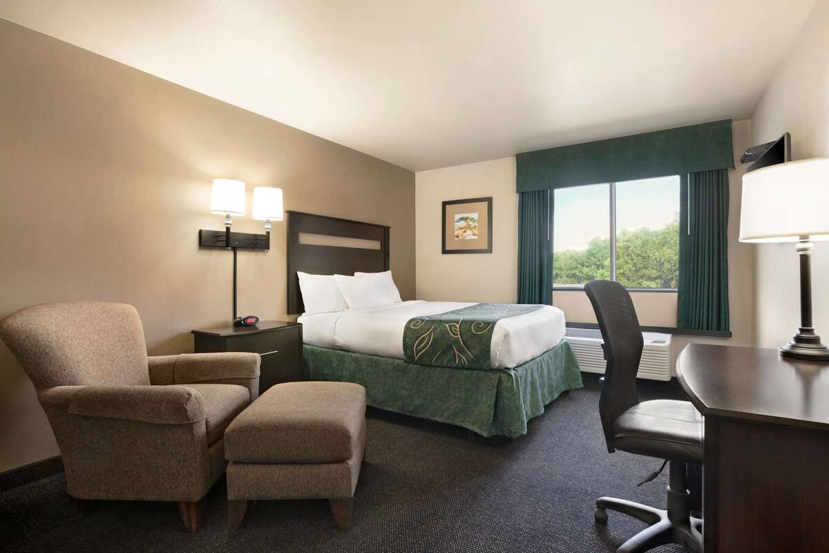 Une chambre d’hôtel avec un lit, deux chaises, un bureau, une lampe et une fenêtre avec des rideaux.