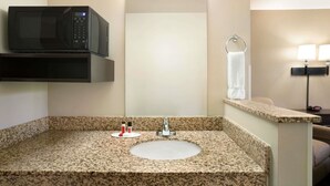 Un baño con lavamanos de encimera de granito, espejo, toallero, microondas y estante pequeño con artículos de aseo.