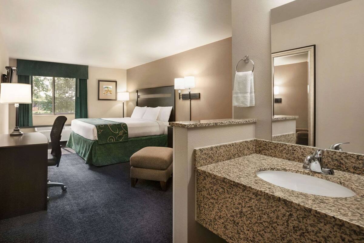 Une chambre d’hôtel comprenant un lit, un bureau, une chaise, une salle de bain avec un lavabo et un miroir, et une fenêtre avec des rideaux.