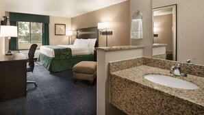Habitación de hotel con cama, escritorio, silla, un pequeño reposapiés, y un baño con lavamanos y espejo de encimera de granito.