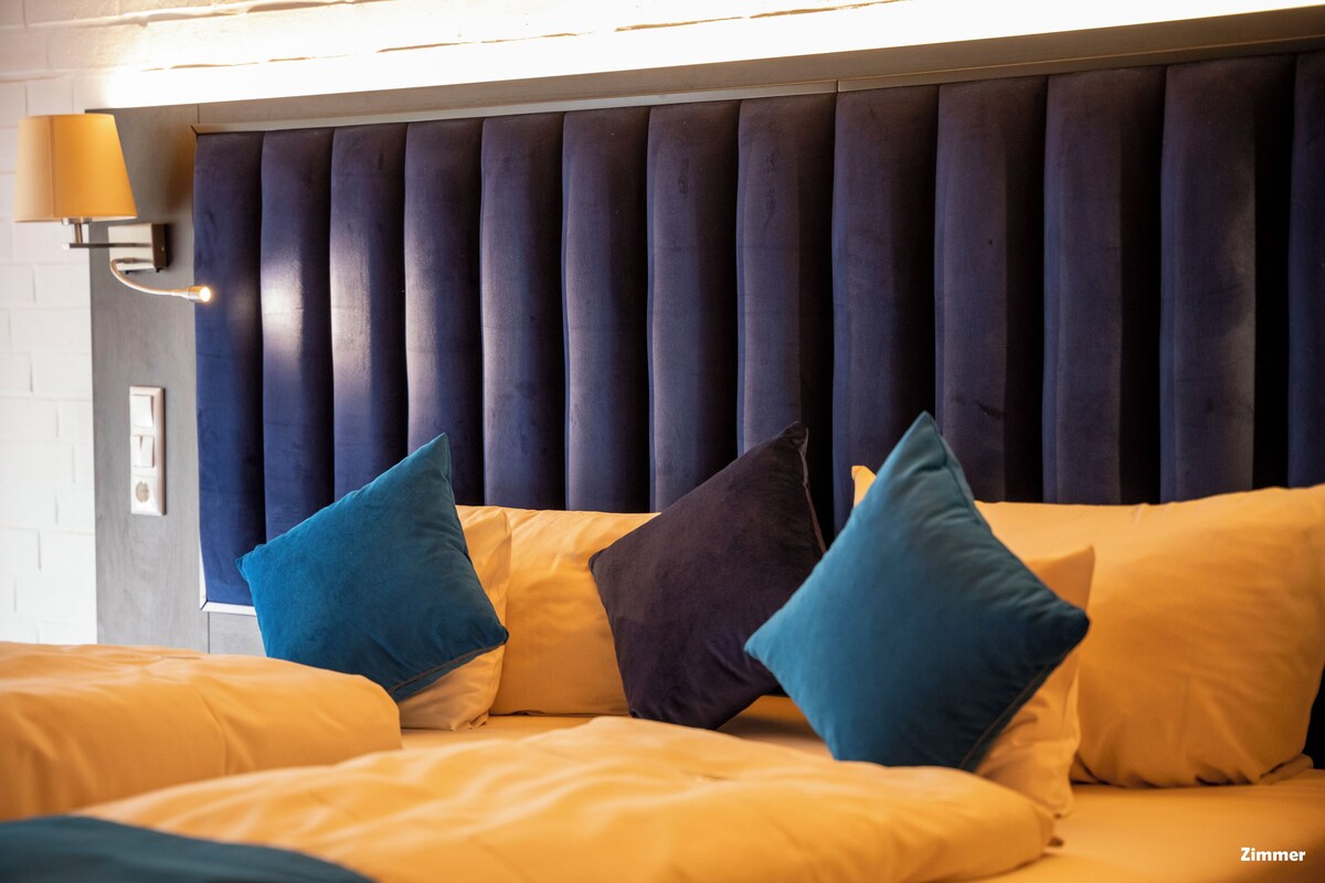 Una habitación de hotel con una cama, un cabecero con paneles verticales y una lámpara fijada a la pared.