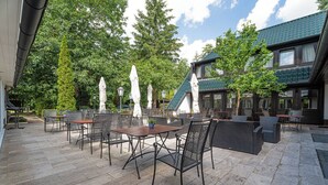 An outdoor patio area with stone flooring, metal chairs, and tables, surrounded by trees and a building with a green roof. - Hotel Zur Heidschnucke (Asendorf)
