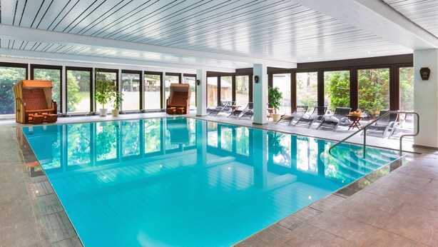 An indoor swimming pool with clear blue water, surrounded by lounge chairs and potted plants. - Hotel Zur Heidschnucke (Asendorf)