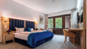 A bedroom with a large bed, a desk, and a chair. There is a window with curtains and a view of greenery outside. - Hotel Zur Heidschnucke (Asendorf)