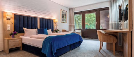 A bedroom with a large bed, a desk, and a chair. There is a window with curtains and a view of greenery outside.