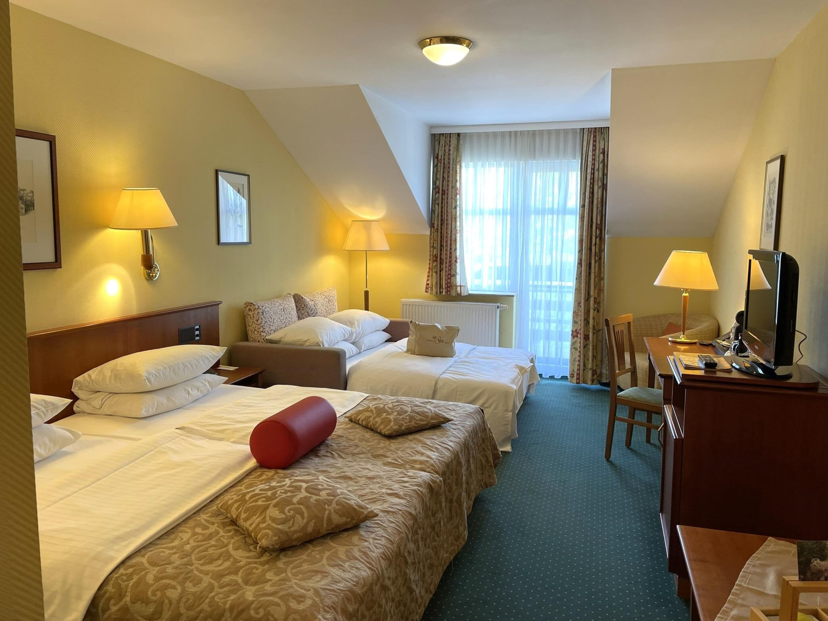 Une chambre d’hôtel avec deux lits, un bureau avec un ordinateur et une chaise. Il y a des lampes de chaque côté du lit et une fenêtre avec des rideaux.