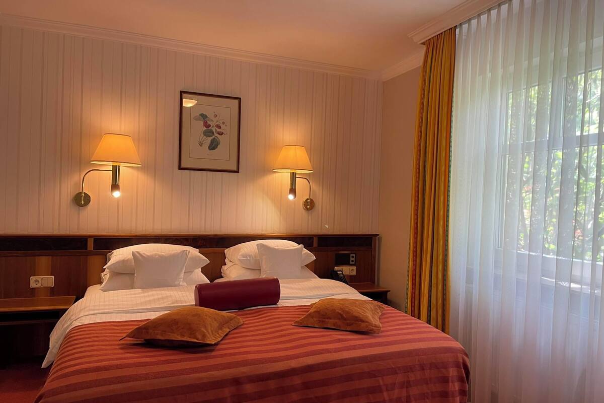 A hotel room with a large bed, two wall-mounted lamps, a framed picture, and a window with curtains.