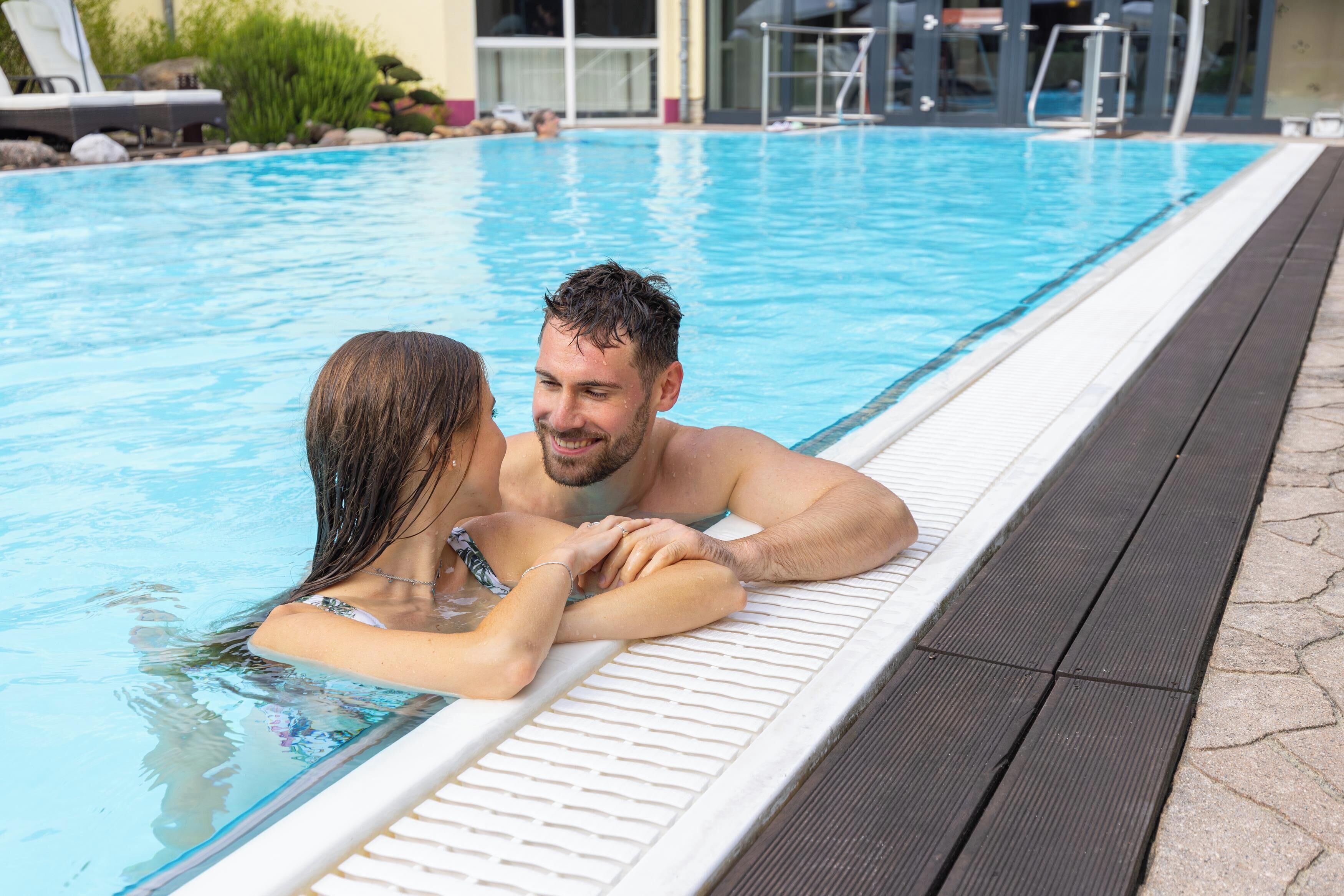 Una pareja en una piscina con borde blanco y cubierta de madera.