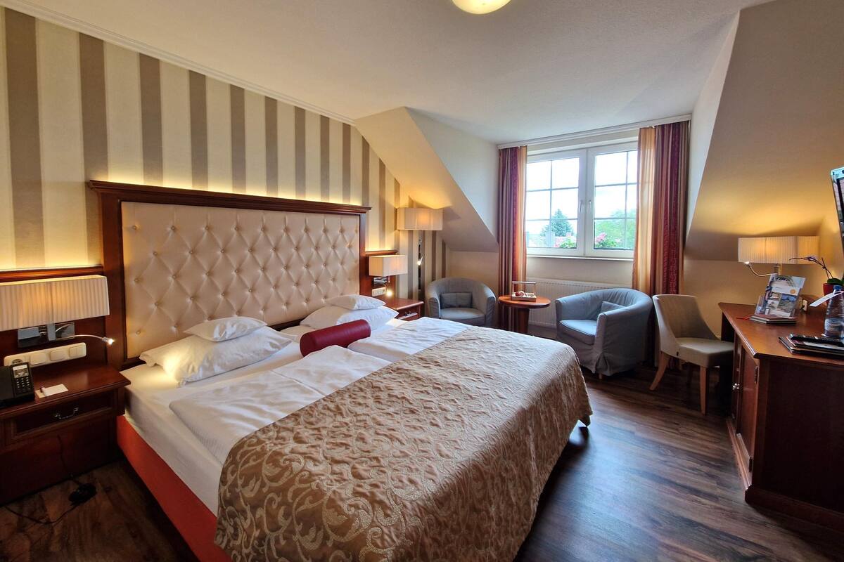 A hotel room with a large bed, a desk, two chairs, a window with curtains, and a ceiling light.