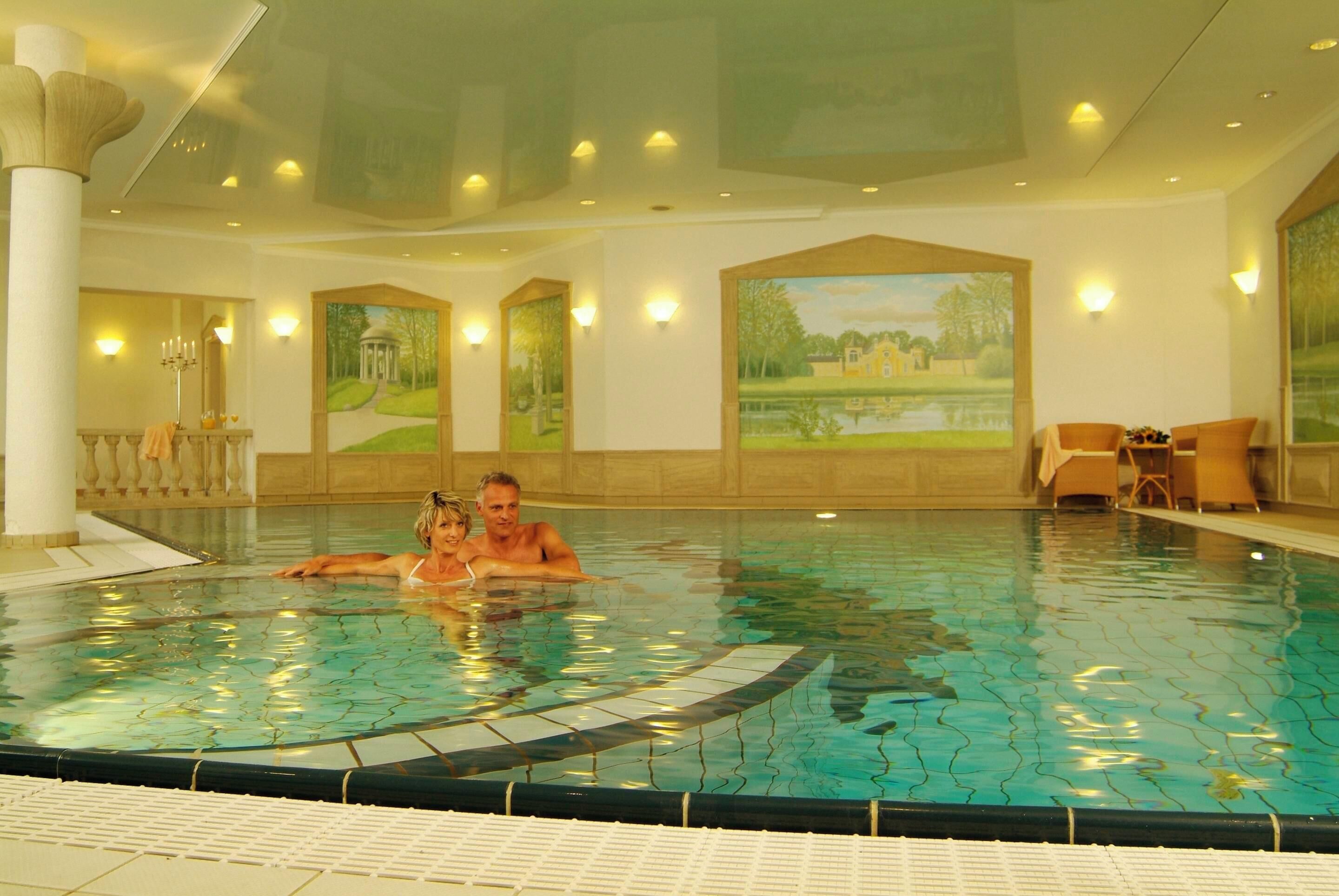 An indoor swimming pool with tiled walls, a central fountain, and a seating area with chairs.