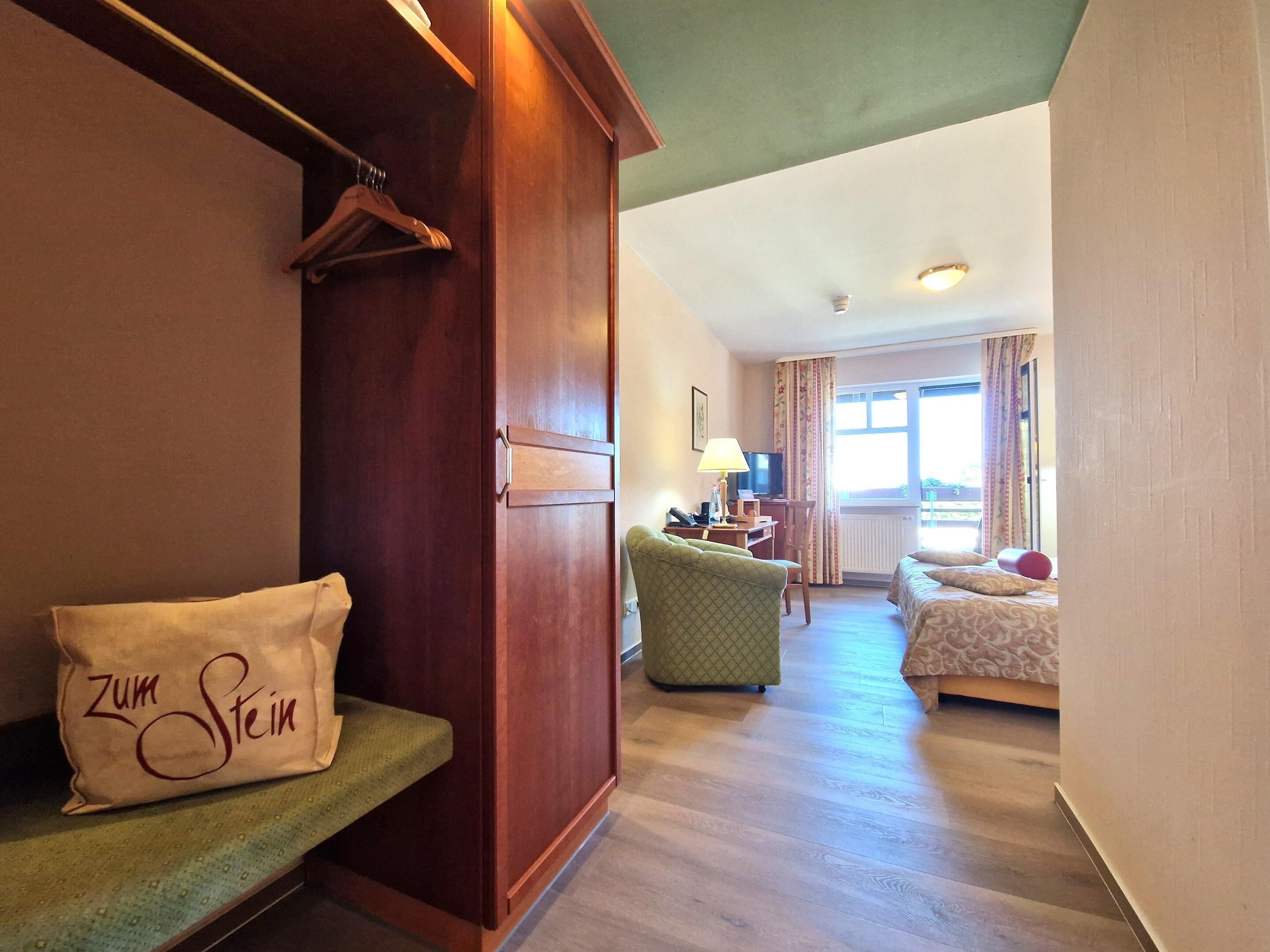 Habitación de hotel con armario de madera, un sillón verde, cama con ropa de cama estampada y una ventana con cortinas.