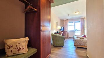 Une chambre d’hôtel avec une armoire en bois, un fauteuil vert, un lit avec une literie à motifs et une fenêtre avec des rideaux.