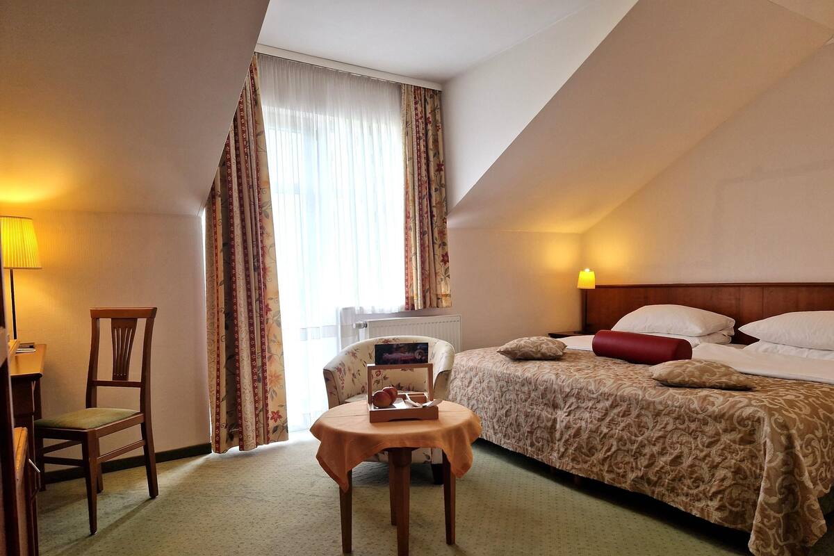 A hotel room with a bed, a chair, a small table, a lamp, and a window with curtains.