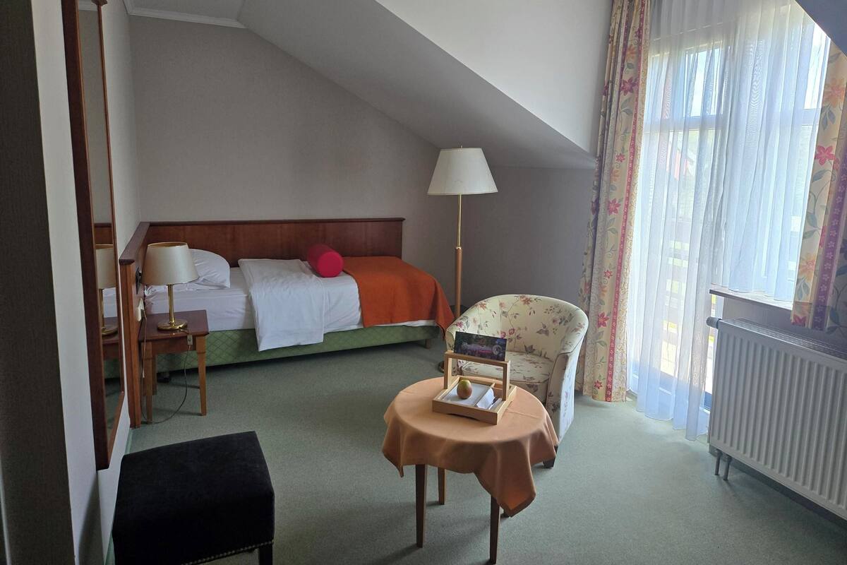 A hotel room with a bed, a chair, a table with a book and an apple, a lamp, and a radiator.