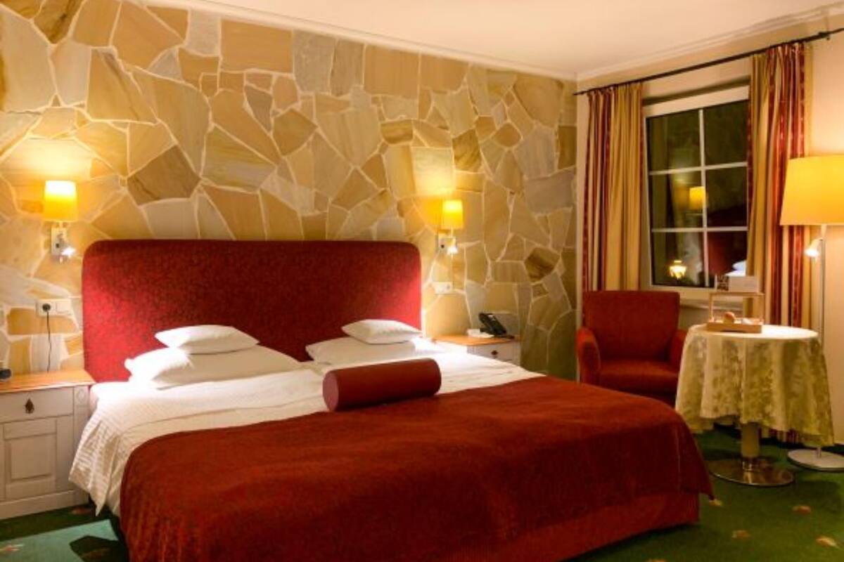 A hotel room with a large bed, stone wall, two bedside lamps, a red chair, and a small table with a lamp.