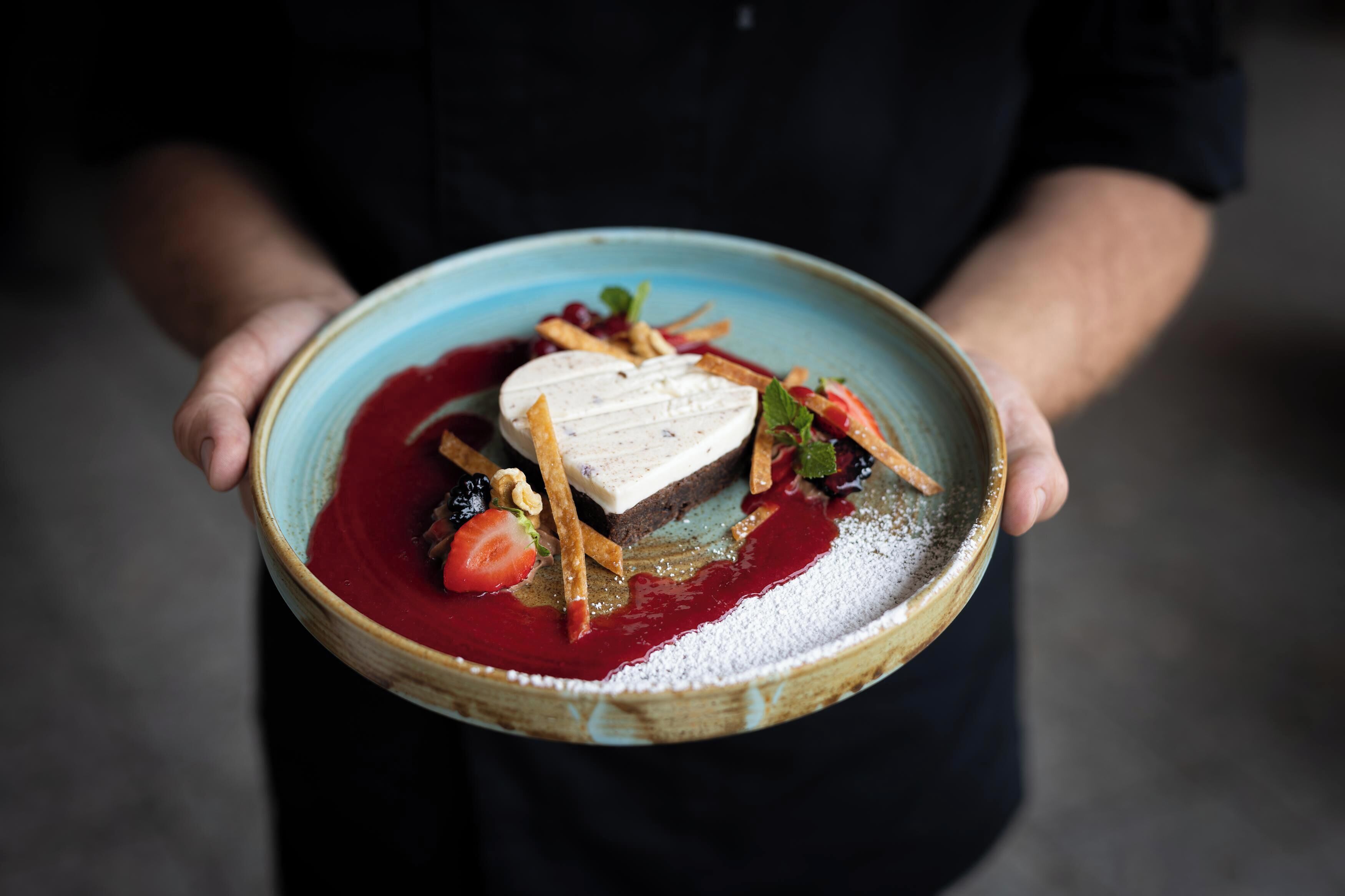 A person holding a plate with a dessert that includes a piece of cake, strawberries, and a red sauce.