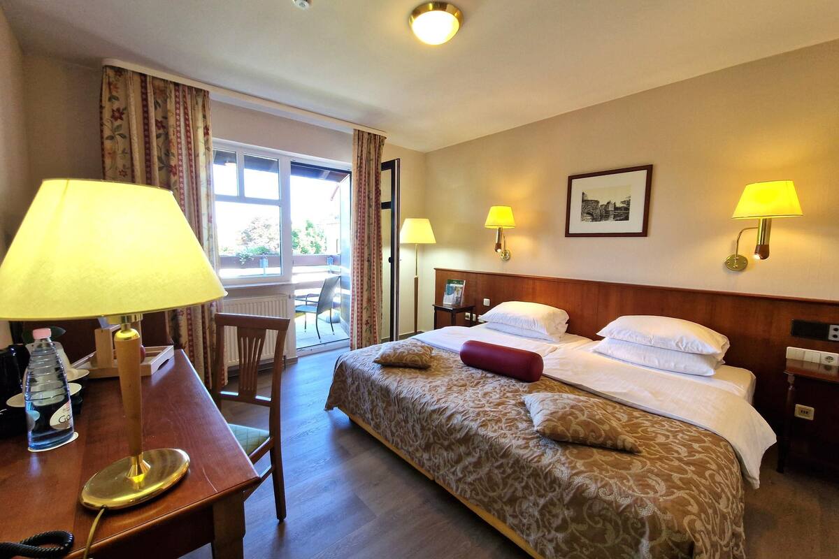 A hotel room with a bed, bedside lamps, a desk with a lamp, a chair, and a window with curtains.