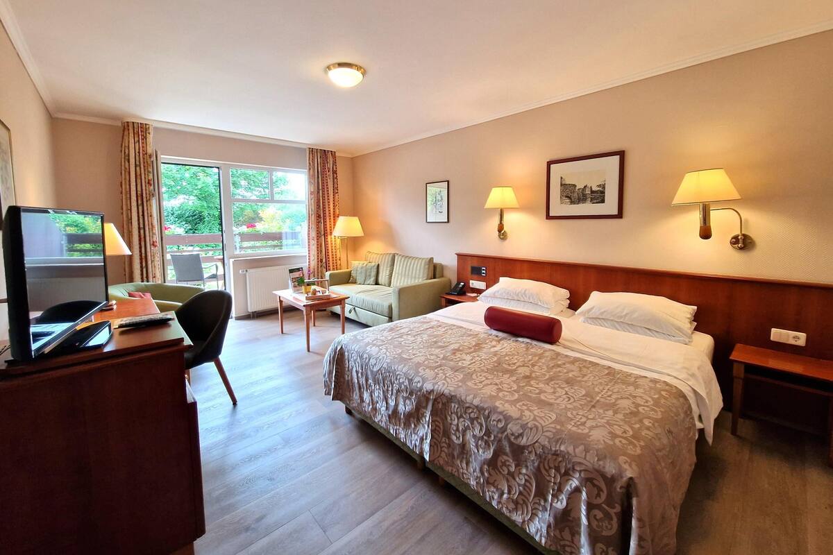 A hotel room with a bed, a desk with a TV, a sofa, and two bedside lamps.