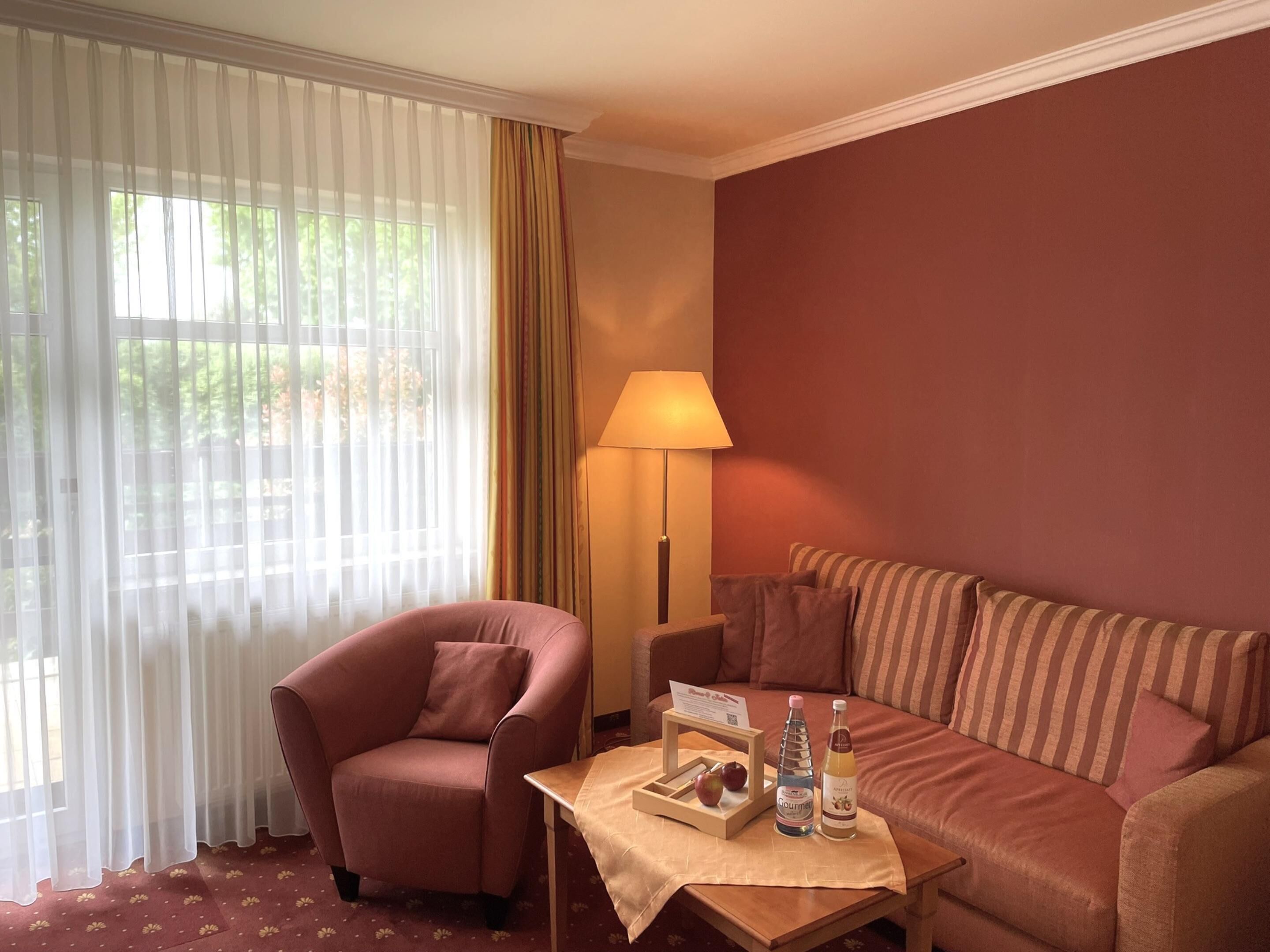 A hotel room with a red sofa, a pink armchair, a table with a tray of apples and bottles, and a lamp.