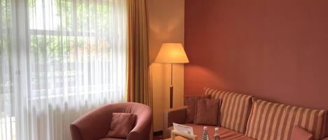 A hotel room with a red sofa, a pink armchair, a table with a tray of apples and bottles, and a lamp.