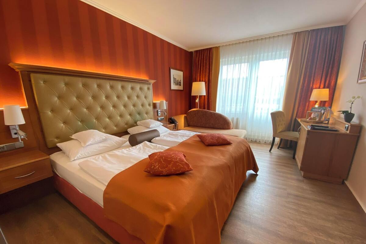 A hotel room with a large bed, a wooden headboard, a desk with a lamp, a chair, and a window with curtains.