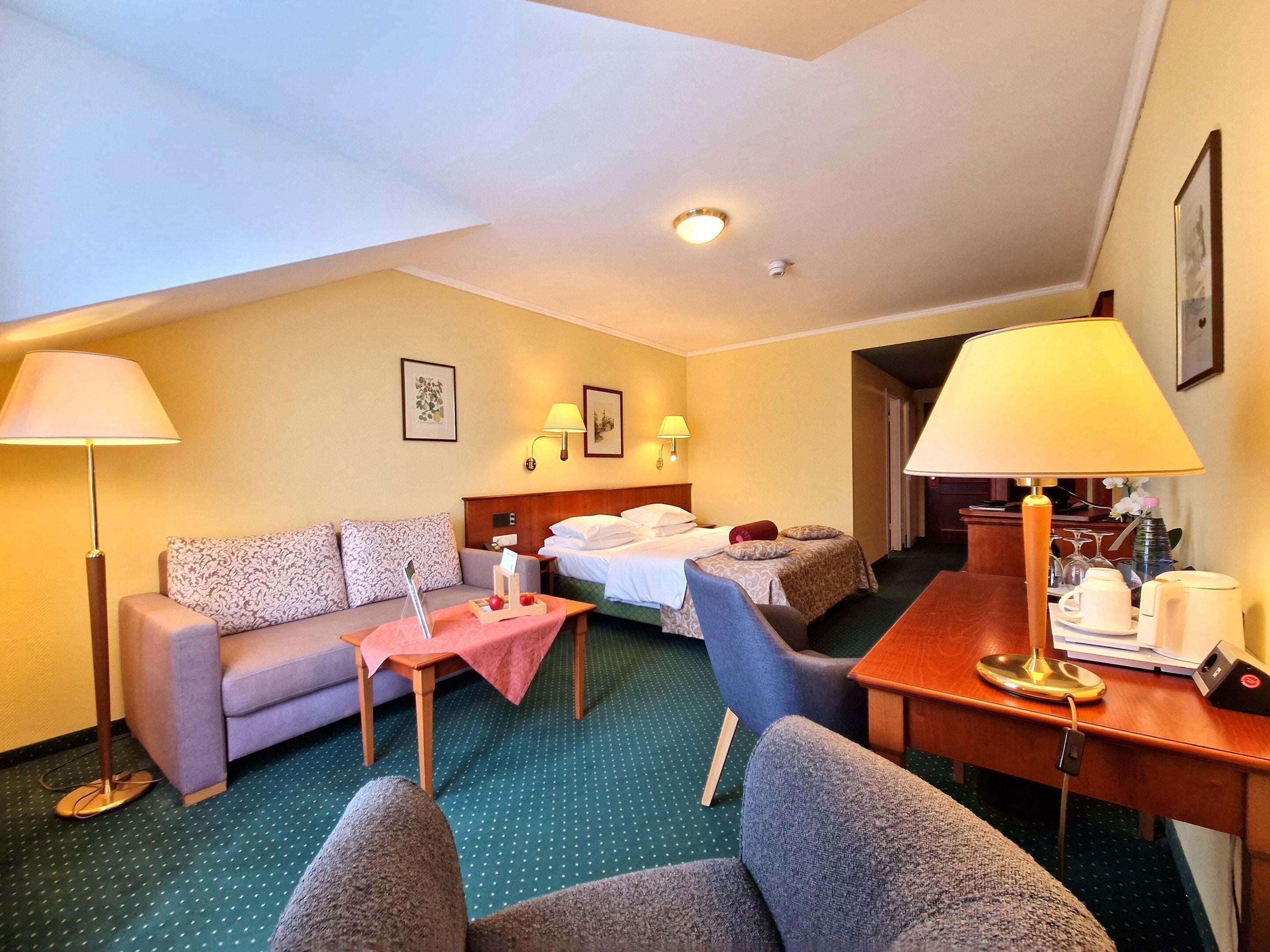 Quarto de hotel com sofá, duas camas de solteiro, uma mesa de jantar com abajur e uma mesinha com toalha de mesa vermelha.