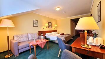 Quarto de hotel com sofá, duas camas de solteiro, uma mesa de jantar com abajur e uma mesinha com toalha de mesa vermelha.