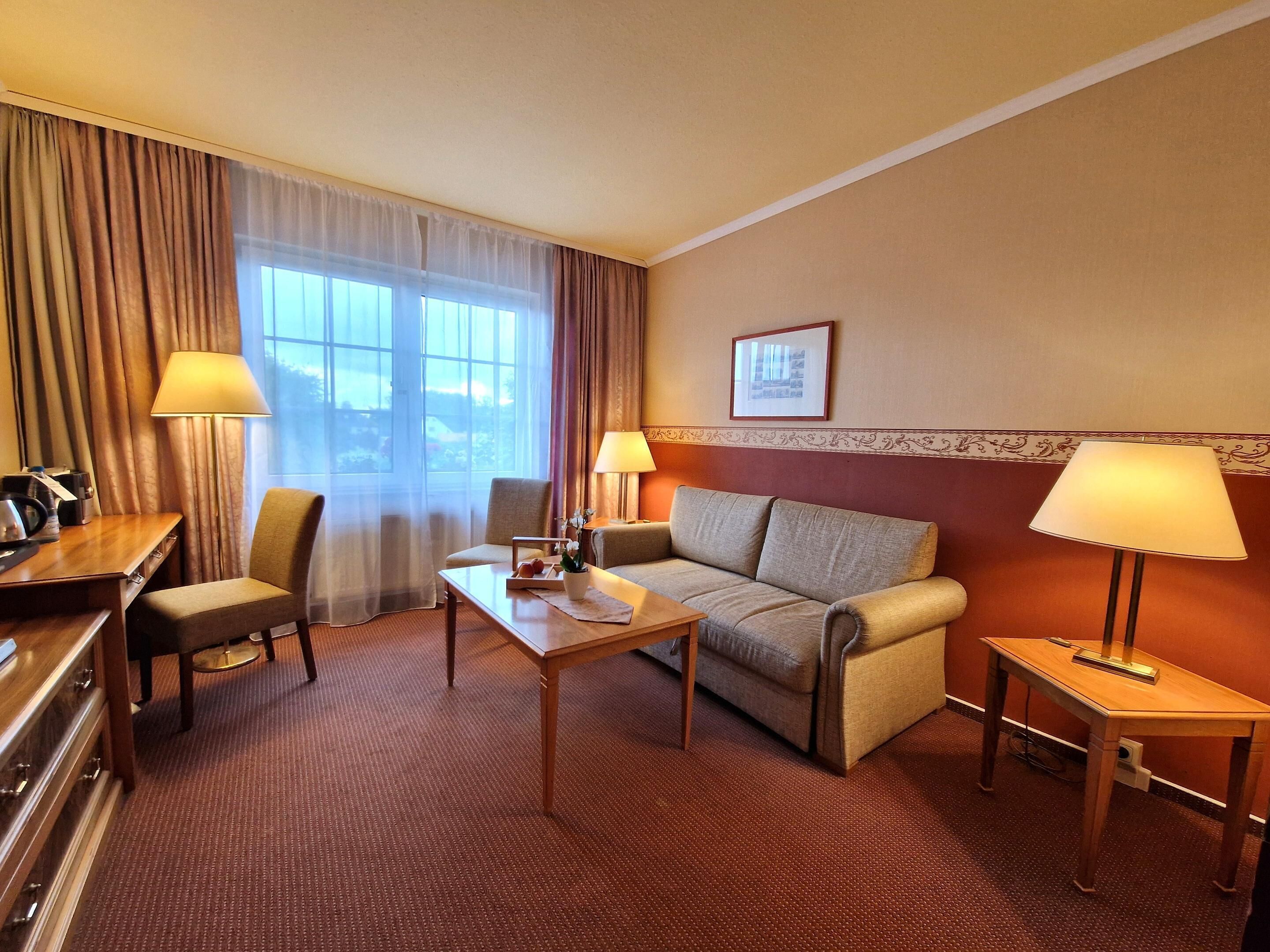 A hotel room with a sofa, two armchairs, a small table, a desk, and a lamp.