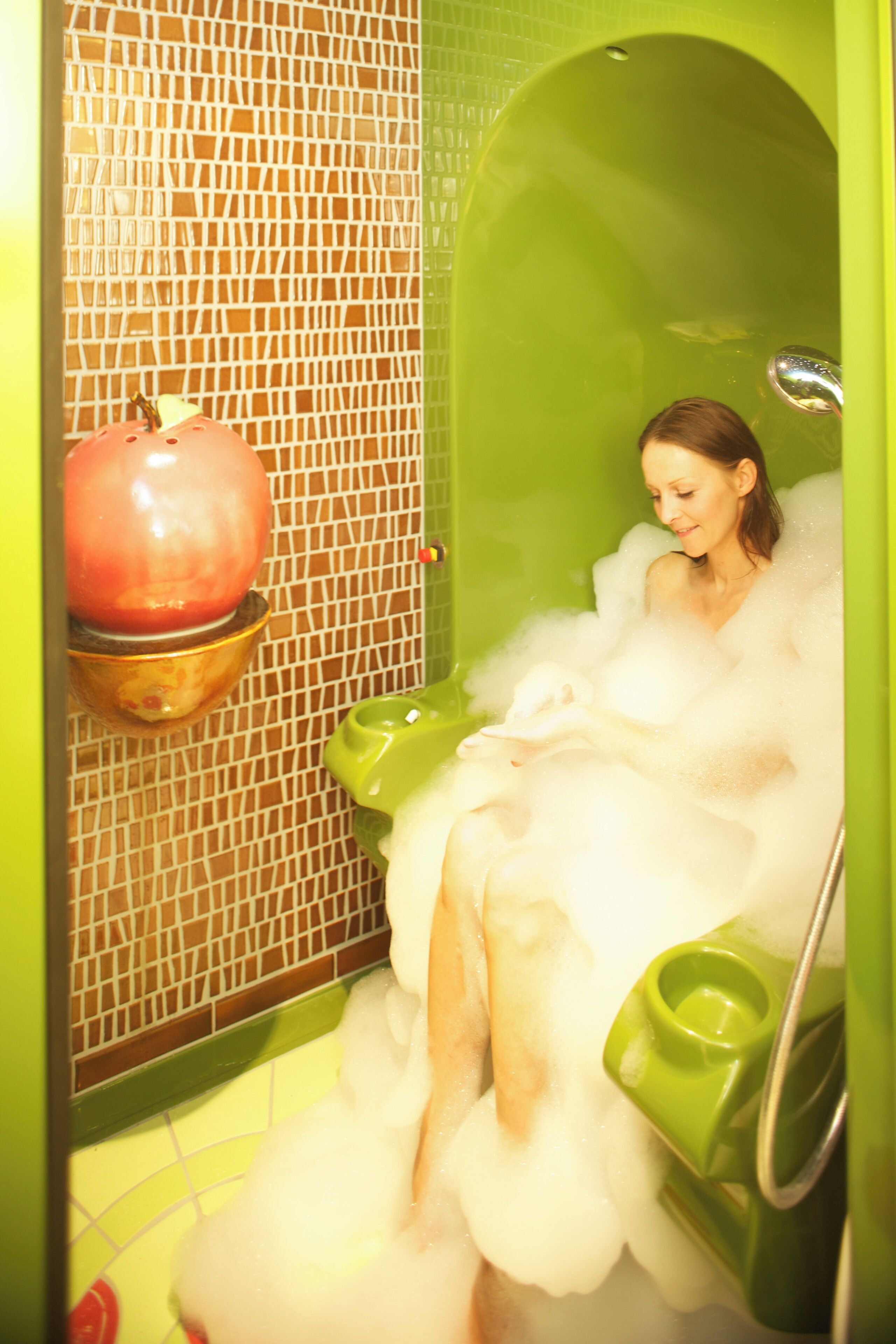 A bathtub filled with bubbles, a green toilet, and a red bowl on the wall.