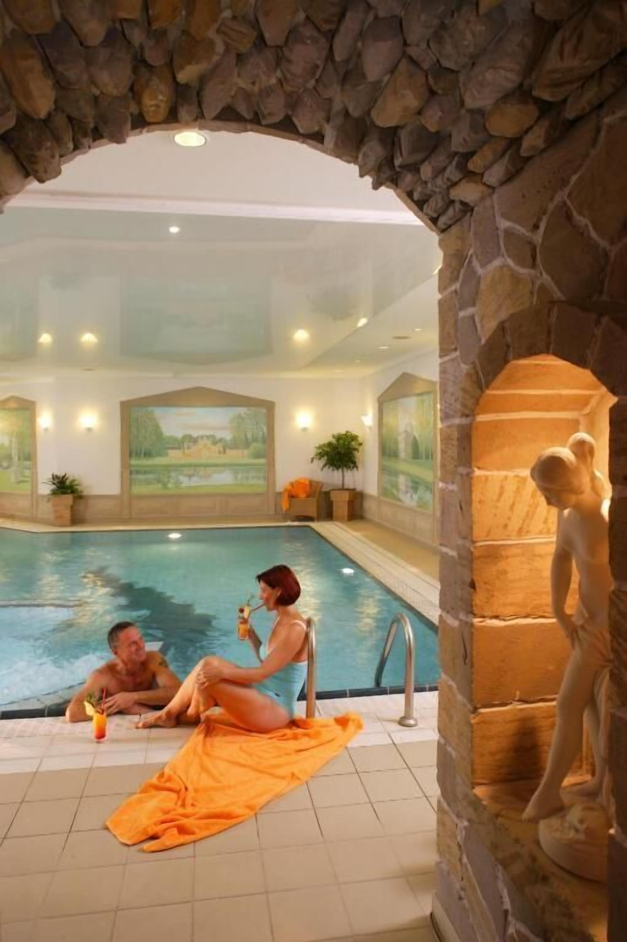 An indoor swimming pool with stone arches, a mural, and a statue.