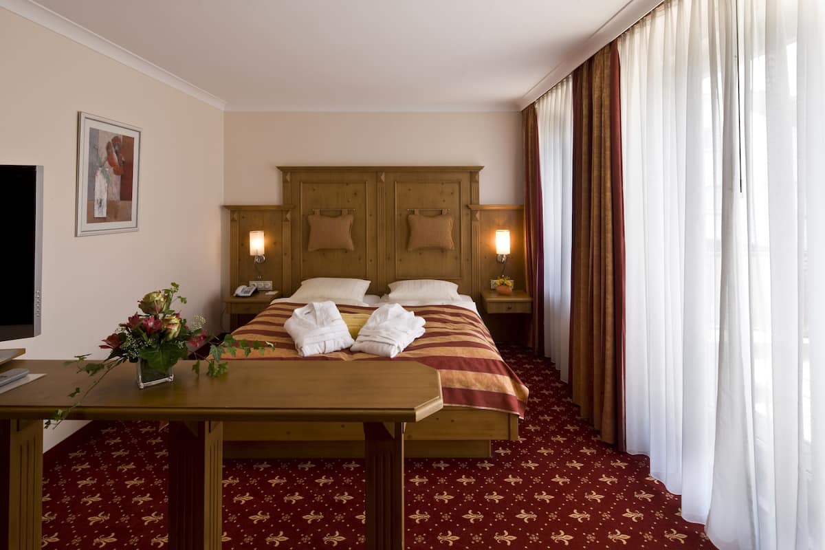 A hotel room with a bed, bedside tables, a TV, a desk with a vase of flowers, and a carpeted floor.